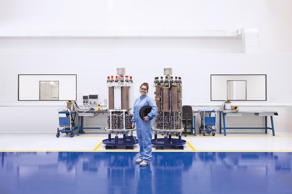 A female OneSubsea employee in a blue jumpsuit stands proudly in a lab in front of two subsea control modules (SCM) in Curitiba Brazil facility.